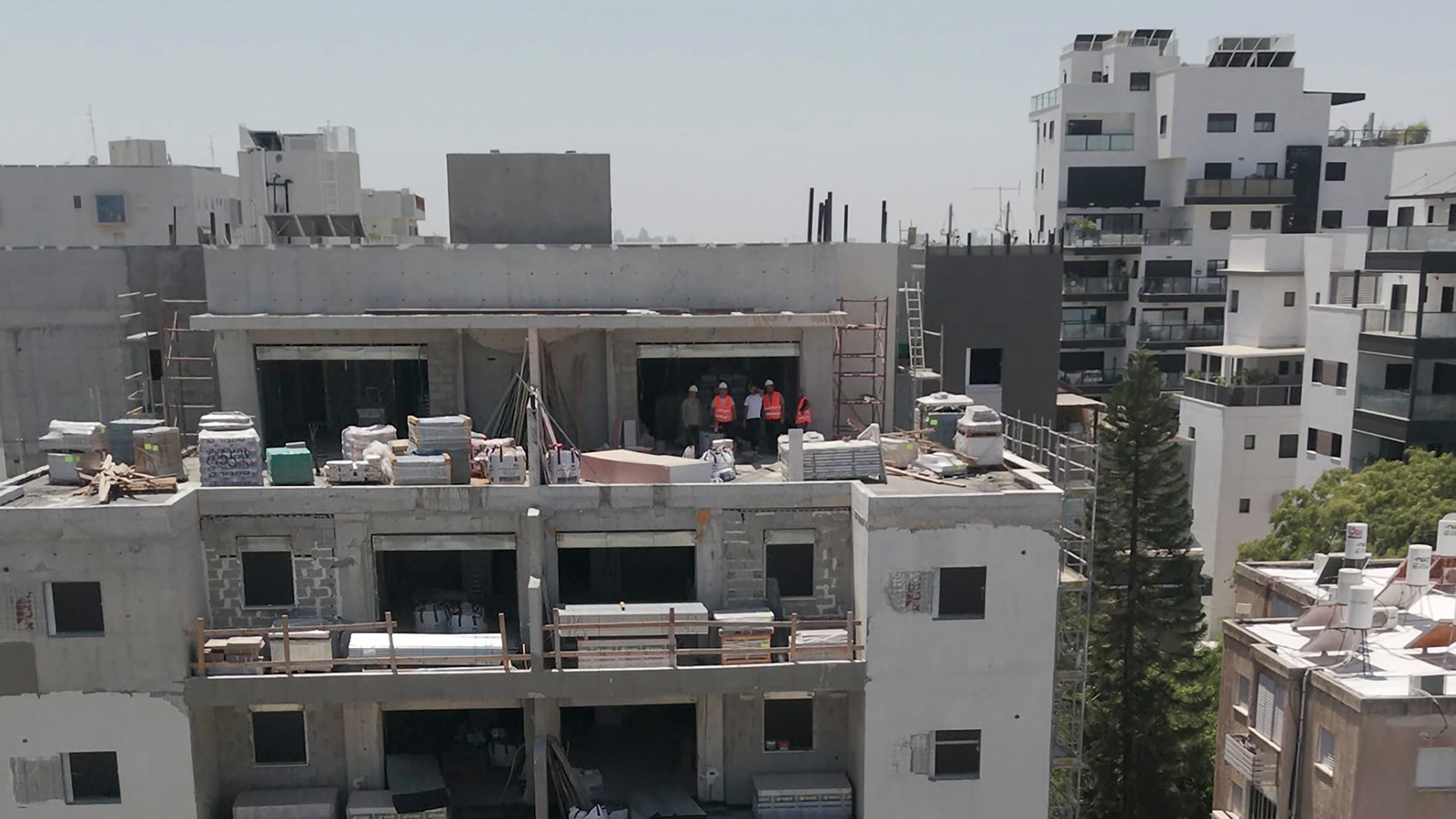 צילום רחפן של בניין מגורים בשלבי בנייה בתוך שכונה עירונית, צוות עובדים קטן נראה בפתח הקומה העליונה